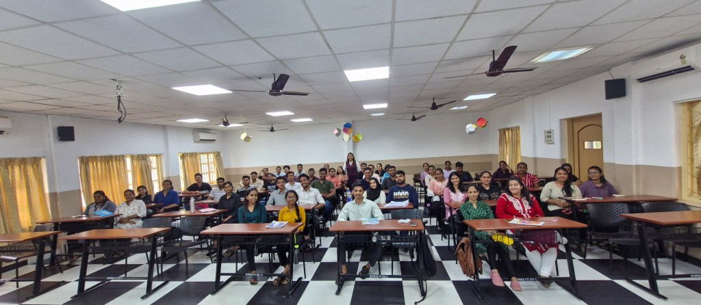 A large group of students sitting at desks in a classroom, with a mix of genders and ethnicities, engaged in learning, in a well-lit environment.