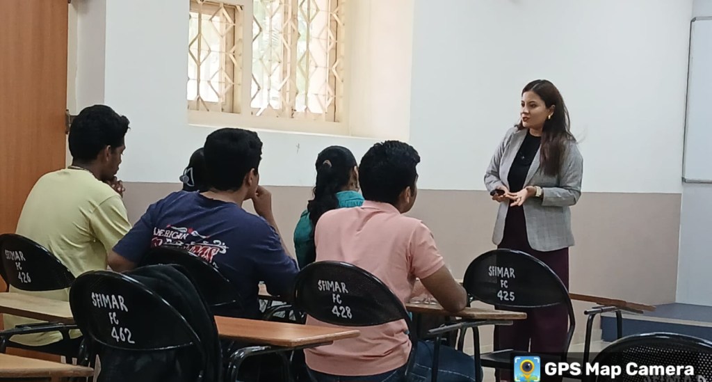 A female instructor speaks to a small group of attentive students seated at desks in a classroom setting.
