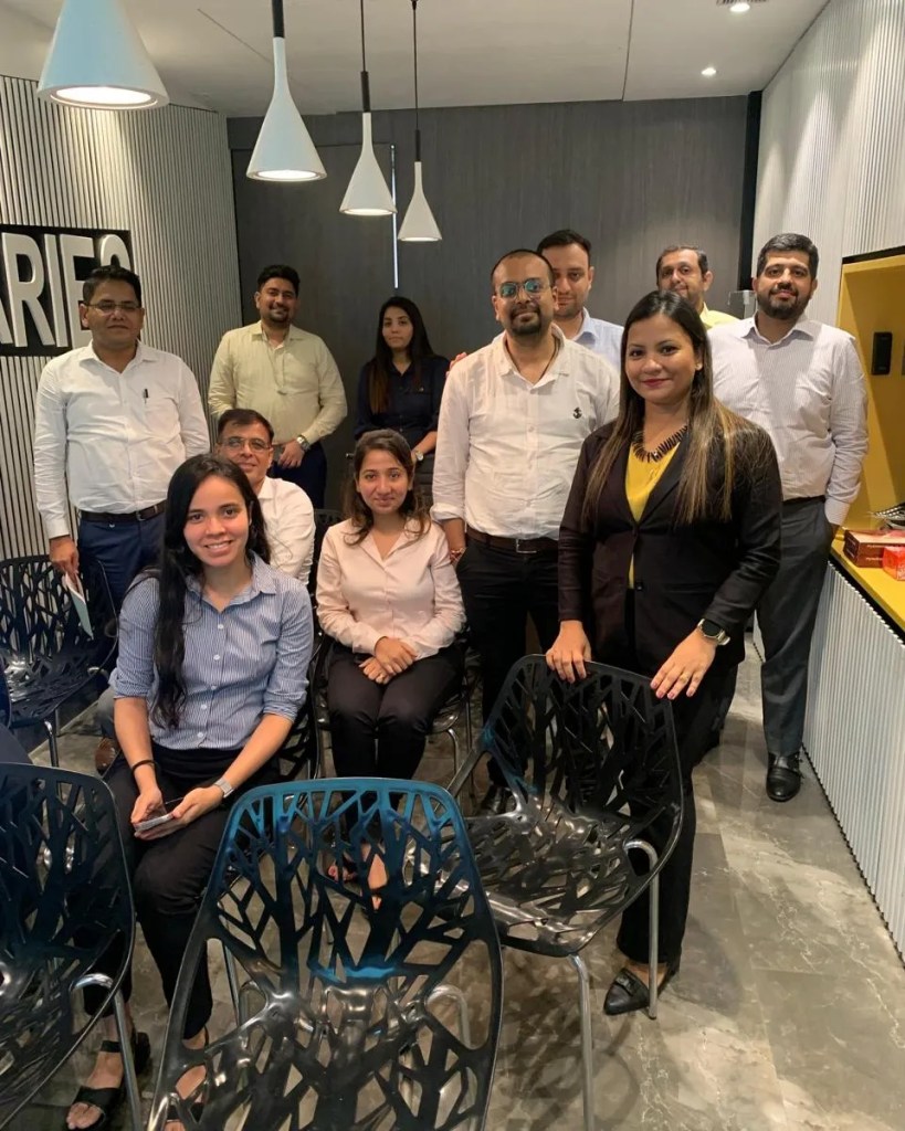 A group of professionals posing together in a modern office setting, with some seated and others standing. The room features stylish lighting and contemporary chairs.
