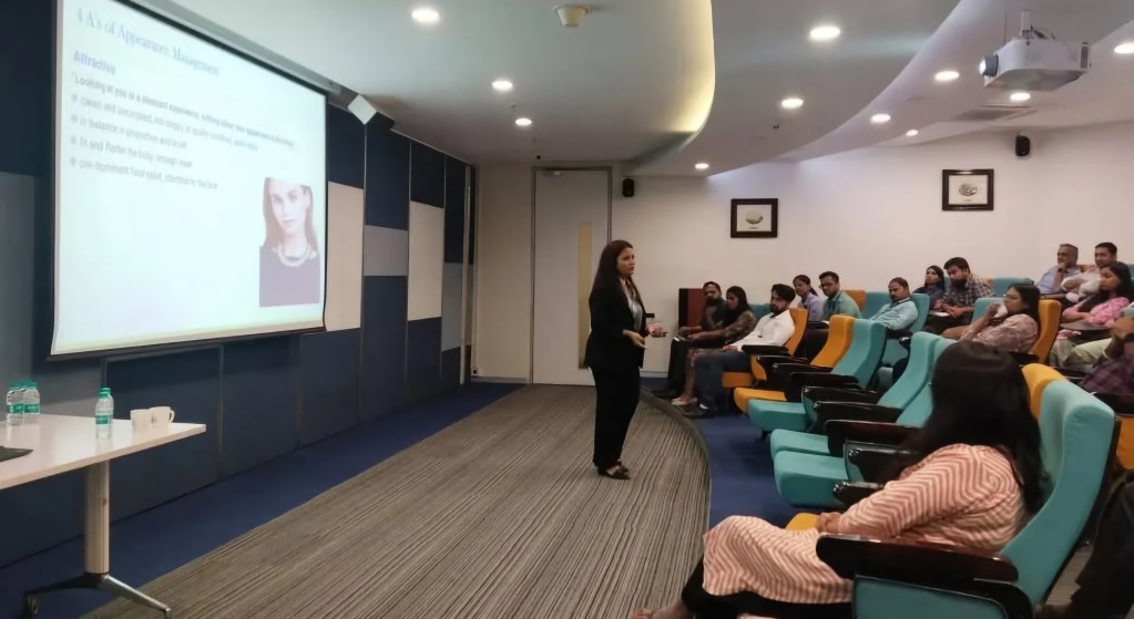 A speaker presenting on appearance management to an audience seated in a semi-circular arrangement. The presentation slide is visible in the background, discussing topics related to attraction. The room features modern seating and a projector.